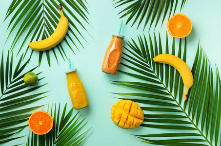 新鲜的橙色, 香蕉, 菠萝, 芒果冰沙和多汁的水果在棕榈叶超过蓝色背景。排毒夏日饮品。素食概念。顶部视图, 平面放置, 复制空间
