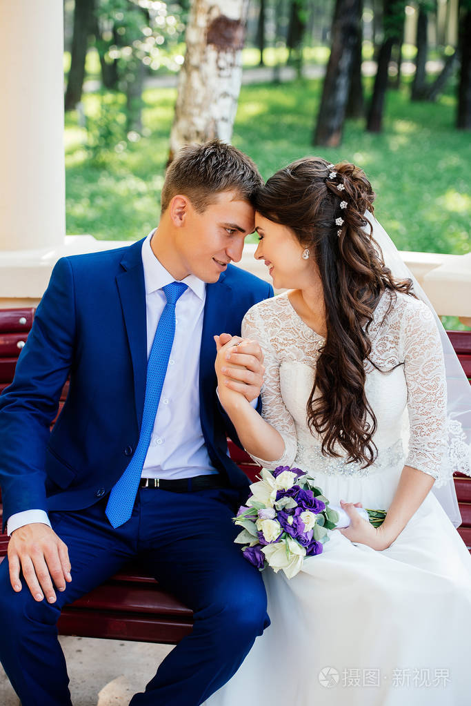 美丽的年轻新娘和新郎坐在长椅上牵着手.婚礼情侣恋爱在婚礼当天