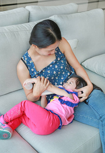 母亲抱着她可爱的亚洲孩子女孩年龄约一岁和九个月，从一瓶在沙发上喝水
