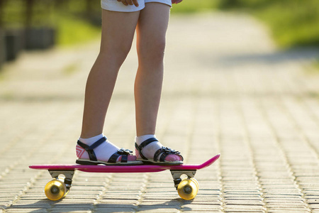 女孩苗条的腿在白色袜子和黑色凉鞋站在人行道上的塑料粉红色滑板上明亮的阳光明媚的夏天模糊的背景。户外活动和健康的生活方式概念图片