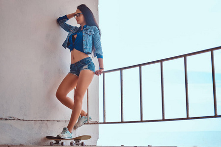 的黑发女孩穿着短裤和牛仔裤夹克站在滑板上, 靠在一个护栏对海岸边