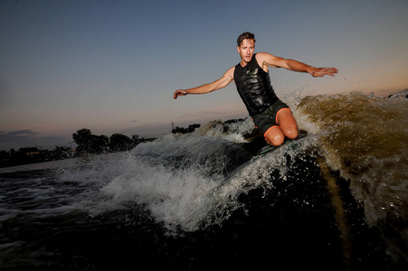 肌肉年轻男子 wakesurfing 在黄昏的背景下, 在美丽的多云的天空波浪