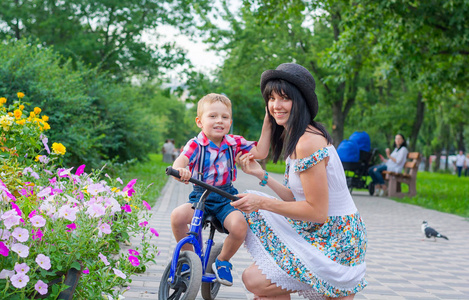 妈妈帮助一个小男孩骑自行车, 在夏日公园里微笑。