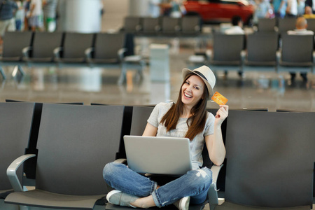 年轻微笑的旅行妇女在帽子坐与交叉的腿, 工作在手提电脑持有信用卡等待在大厅大厅在机场。周末出游的旅客出国旅行。航空飞行概念