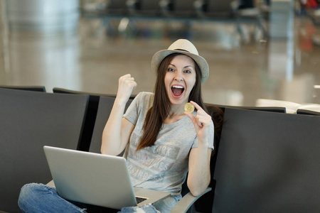 年轻的狂喜的旅行妇女在帽子坐在笔记本电脑持有比特币, 做赢家的手势, 在大堂大厅在机场等待。周末旅客出国旅行。航空飞行概念
