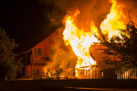 晚上烧木房子。明亮的橙色火焰和浓密的烟雾下的瓷砖屋顶在黑暗的天空, 树木剪影和住宅邻居平房的背景。灾害和危险概念