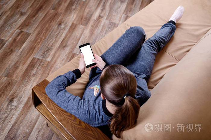 美丽的年轻女子坐在沙发上,使用智能手机和短信.在家里的沙发上休息.