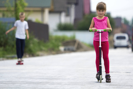 可爱的年轻金发女孩在粉红色的衣服上的滑板车和英俊的男孩在阳光明媚的街道上玩在模糊的夏天明亮的背景。儿童活动, 游戏和有趣的概念