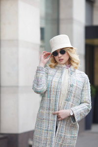 一个美丽的时尚金发女人穿着大衣, 帽子和太阳镜在城市街道上