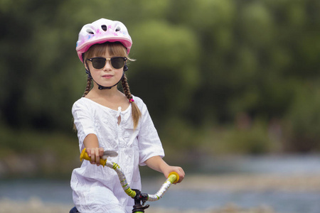 特写肖像的骄傲漂亮的年轻女孩在白色的衣服, 太阳镜与长金色的辫子穿着粉红色安全头盔骑儿童自行车在模糊的绿树夏日复制空间背景