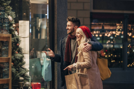 一对夫妇在圣诞节做一些橱窗购物的侧面视图。中成年男性有他的手臂周围的中年女性, 他们正在谈论广域在窗口