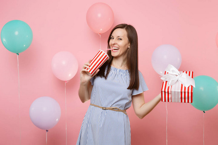 蓝色礼服的快乐的笑的妇女的肖像持有红色框与礼物从塑料杯在柔和的粉红色背景与五颜六色的空气气球喝苏打或可乐。生日聚会