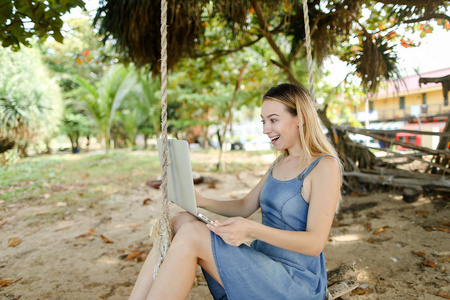 年轻快乐的女孩使用笔记本电脑和骑在沙滩上摇摆