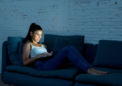 年轻迷人的西班牙裔和放松的女人在家里微笑快乐的沙发沙发上使用笔记本电脑在黑暗的黄昏光享受观看在线电影或沉迷于网络社交网络