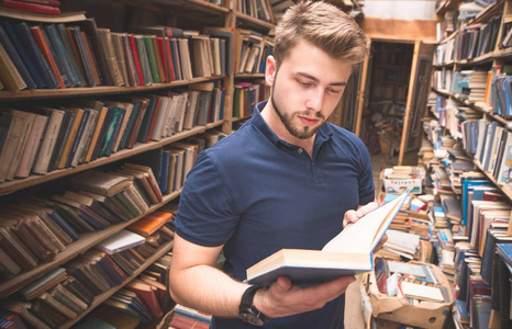 有胡子的帅哥站在旧图书馆里, 手里拿着书看书.