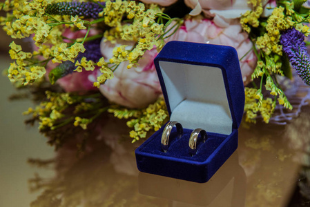 桌子上有两个结婚戒指在蓝色的地壳上。婚礼当天美丽的粉红色牡丹花新娘花束
