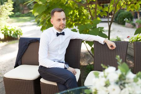 一个英俊的年轻商人穿着白衬衣, 在商务晚宴上绑一只蝴蝶, 等待他的同事独自在一张夏日餐桌上用餐。