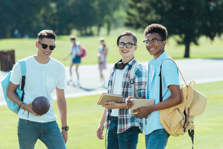 多民族青少年男孩与书和背包微笑着在公园的相机