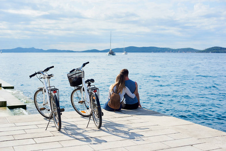 游客夫妇的后视图, 男子和女孩与背包坐在自行车上的高铺石人行道上观看漂浮游船在清澈的蓝色海水。活动假日概念