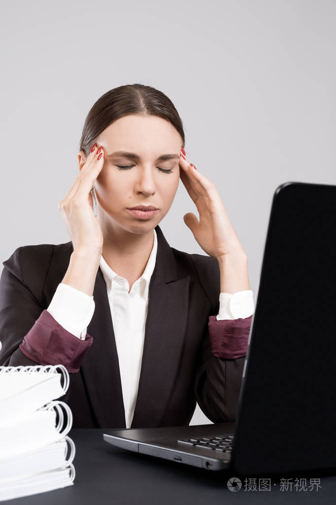 年轻疲惫的商业妇女坐在办公桌前的工作暂停与笔记本电脑的文件和书籍