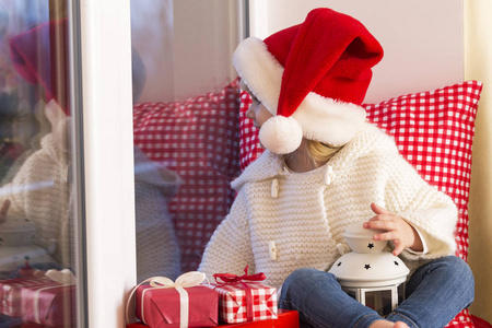 幸福的家庭。一个小女孩在圣诞老人的帽子坐在窗台上的窗口和等待圣人