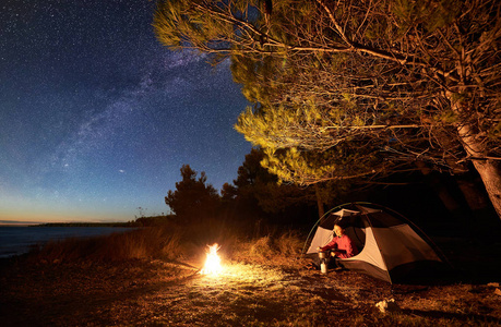 晚上在湖边露营。快乐的女性游客休息在帐篷入口处, 在篝火旁的大树下, 在星空和银河下的燃气燃烧器上准备食物, 享受清澈湛蓝的海水