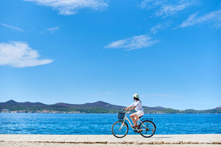 女孩骑单车在白色夏天关闭和帽子骑自行车沿石人行道上蓝色波光粼粼的