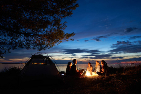 晚上夏天在湖岸边露营。一群五年轻的游客坐在海滩周围的篝火附近的帐篷在美丽的蓝色傍晚的天空下。旅游友谊与自然之美