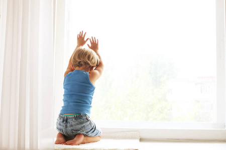 孩子失去了母亲的观念。小可爱的金发碧眼的三岁男孩坐在窗台上触摸玻璃窗, 看着伤心。在蓝色坦克顶部的孩子独自玩。背景, 特写, 复