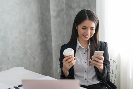 亚洲商业女孩在她的工作站举行咖啡杯和智能手机, 而微笑