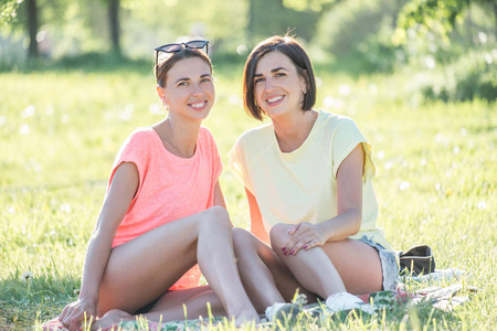 两个欢快的女孩坐在草坪上的户外肖像和愉快的日子一起在公园在晴天