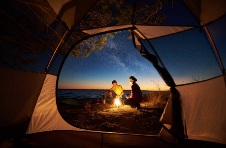 日落时在海边露营, 从旅游帐篷里查看。年轻的旅游情侣, 男人和女人准备食物的燃气燃烧器, 坐在篝火旁的蓝色海水和星空背景