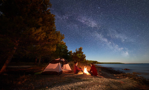 晚上在岸上露营。男子和女子徒步旅行者在帐篷前休息, 在黄昏的天空中充满了星星和银河的蓝色水和森林背景下的篝火。户外生活方式概念