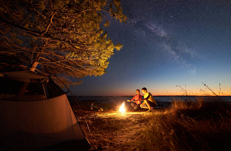 夜帐篷露营在湖在蓝色星空下。年轻的旅游者, 男人和女人在篝火上热身在清澈的水和城市的灯光在遥远的岸边地平线背景。旅游与爱情理念