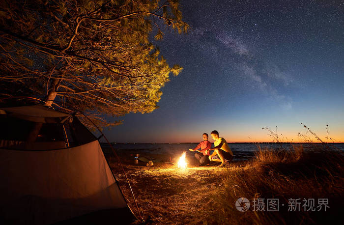 年轻的旅游者, 男人和女人在篝火上热身在清澈的水和城市的灯光在遥远