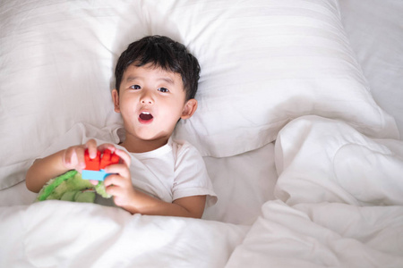 3岁小可爱的亚洲男孩在家里的白色衬衫在床上, 孩子躺在玩和微笑的白色床上枕头和毯子, 顶部视图与复制空间添加文本