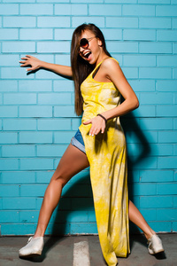 年轻漂亮的女孩摆姿势和微笑附近蓝色墙背景中黄色的裙子 短裤 衬衫 太阳镜