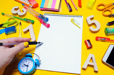 学生在笔记本上做笔记。复制空间。在一个黄色背景的办公桌上的学校配件。教育的概念。文具。手表, 彩色钢笔, 电话, 标记。从上面查
