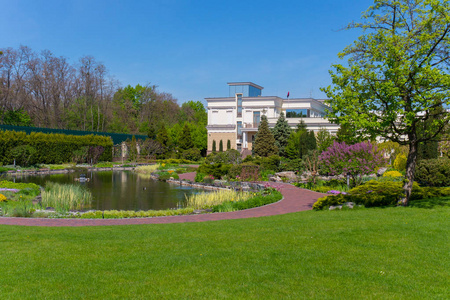 一栋两层的白色现代豪宅, 靠近绿色的草坪上, 装饰着池塘, 在蓝色晴朗的天空下保存着花坛和树木。
