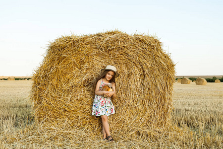 可爱的女孩戴着草帽, 在温暖阳光明媚的夏日傍晚, 在麦田里快乐地散步。可爱的小孩子在黑麦领域在日落与一篮子面包