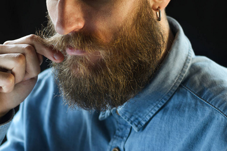 穿着蓝色牛仔衬衫的大胡子男子用手指捻着胡子。黑色背景特写的时髦人物肖像