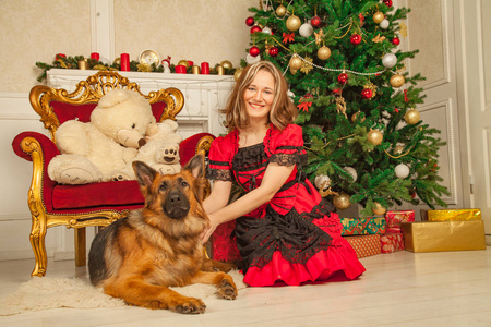 美丽的女孩在红色神话般的礼服摆在她的牧羊人大狗和美味的馅饼在她的米色房间的背景下的圣诞树