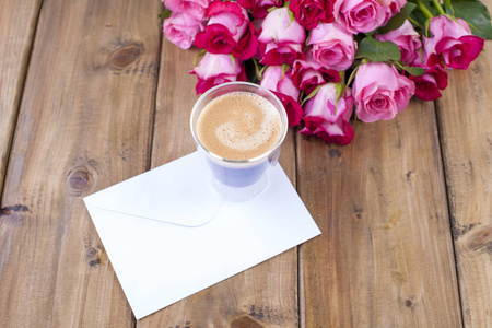 一束新鲜的粉红色玫瑰和一杯浓咖啡。木质背景。文本或明信片的可用空间。用于书写的白色信封