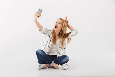 欢快的年轻休闲女孩的肖像坐在自拍与移动手机的腿交叉孤立的白色背景