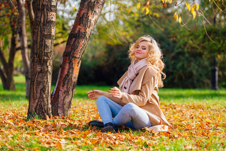 年轻美丽的女人坐在地上的树下覆盖秋天的黄叶