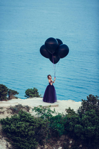 在持有气球对天空和大海的黑连衣裙年轻时尚美女