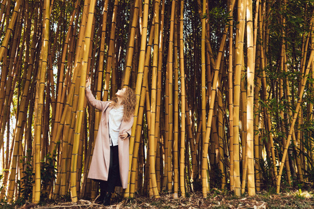 一个年轻的好奇女孩穿着长粉红色的大衣, 享受自然和高竹树