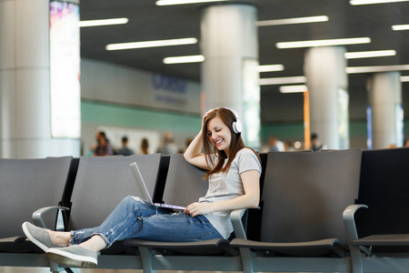 年轻美丽的旅行妇女与耳机收听音乐在笔记本电脑上, 等待在大堂大厅在国际机场。周末出游的旅客出国旅行。航空飞行概念