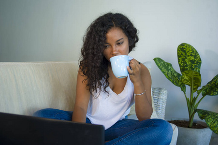 年轻有吸引力和宽松的黑人美国学生的生活方式肖像坐在家里沙发沙发与笔记本电脑一起喝咖啡或茶穿牛仔裤和单线
