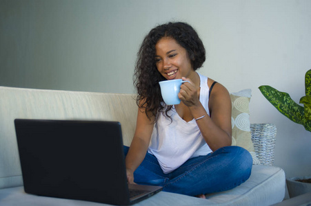 年轻有吸引力和轻松的黑人美国学生的生活方式肖像坐在家里沙发沙发与笔记本电脑一起喝咖啡或喝茶穿牛仔裤